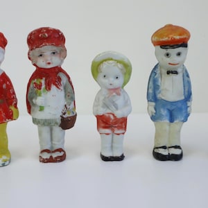 May include: Five antique bisque dolls are displayed. The dolls are dressed in colourful outfits, including red, blue, and yellow. Each doll has painted facial features and is wearing a hat. The dolls are standing on a white surface.