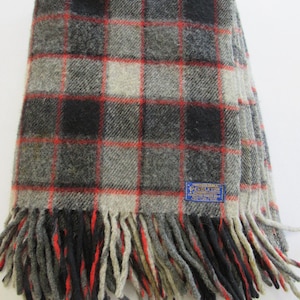 May include: A folded wool blanket with a plaid pattern in shades of gray, black, and red. The blanket has a fringed edge and a blue label with white text. The blanket is a cozy home textile.