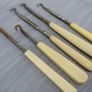 May include: A set of four button hooks featuring long, cream-colored handles and metallic shafts with curved hooks at the ends. The metal components show signs of oxidation and age, while the handles have a smooth, elongated, and tapered shape.