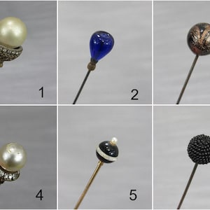 May include: A collection of six hat pins featuring diverse decorative tops: a pearl in a crystal cup, a blue glass teardrop, a black and gold patterned sphere, a pearl in a crystal band, a black and white striped bead, and a textured black sphere.