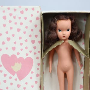 May include: A vintage doll with brown hair and blue eyes, presented in a box adorned with pink hearts. The doll is nude and has a delicate, vintage appearance. The box features a double heart design.