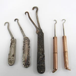 May include: A collection of five antique button hooks with varied designs. Three feature ornate silver-toned metal handles with intricate engravings, while two have slender, textured copper-toned cylindrical handles with small hooks at the ends.