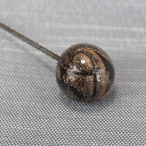 May include: A hat pin featuring a spherical head with a swirling pattern of black and metallic bronze glitter. The round bead is attached to a long, thin, straight metal pin that extends diagonally from the sphere toward the upper left corner.