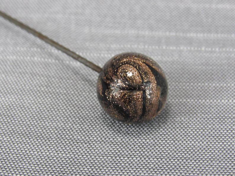 May include: A hat pin featuring a spherical head with a swirling pattern of black and metallic bronze glitter. The round bead is attached to a long, thin, straight metal pin that extends diagonally from the sphere toward the upper left corner.