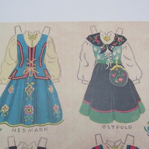 Puede incluir: Ilustraci&oacute;n de dos mu&ntilde;ecas de papel con trajes folcl&oacute;ricos tradicionales. La primera es azul con detalles florales y el texto "HED MARK". La segunda es negra y verde con un bolso floral y el texto "&Oslash;STFOLD".