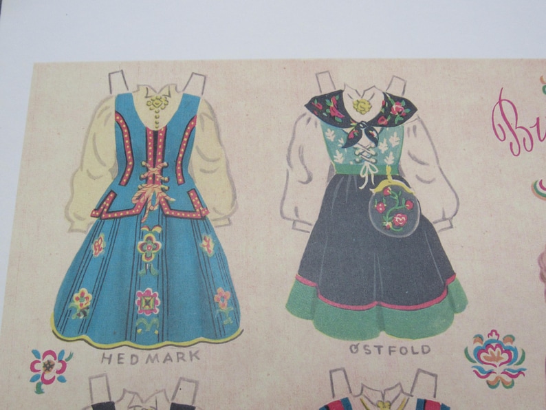 Puede incluir: Ilustraci&oacute;n de dos mu&ntilde;ecas de papel con trajes folcl&oacute;ricos tradicionales. La primera es azul con detalles florales y el texto "HED MARK". La segunda es negra y verde con un bolso floral y el texto "&Oslash;STFOLD".