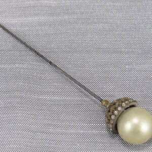 May include: A long, slender metal hat pin featuring a spherical, cream-colored faux pearl at the end. The base of the pearl is set within a small, dome-shaped metal cap encrusted with tiny, clear, sparkling rhinestones that catch the light.