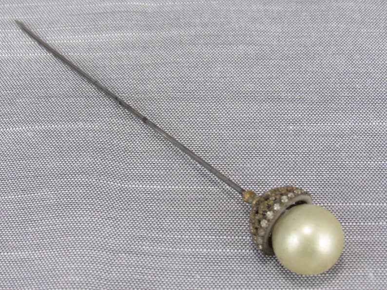 May include: A long, slender metal hat pin featuring a spherical, cream-colored faux pearl at the end. The base of the pearl is set within a small, dome-shaped metal cap encrusted with tiny, clear, sparkling rhinestones that catch the light.