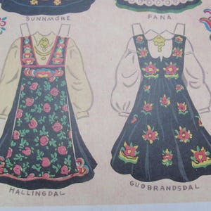 Puede incluir: Ilustraci&oacute;n de dos vestidos tradicionales. Los vestidos son de color azul oscuro con estampados florales en rojo, rosa y amarillo. El vestido de la izquierda est&aacute; etiquetado como "HALLINGDAL" y "SUNNM&Oslash;RE". El vestido de la derecha est&aacute; etiquetado como "GUDBRANDSDAL" y "FANA".
