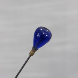 May include: A straight pin featuring a teardrop-shaped cobalt blue glass head attached to a thin metal shaft with a small brass-colored collar. The glass head has a smooth, glossy finish and is securely fastened to the pointed metal needle.