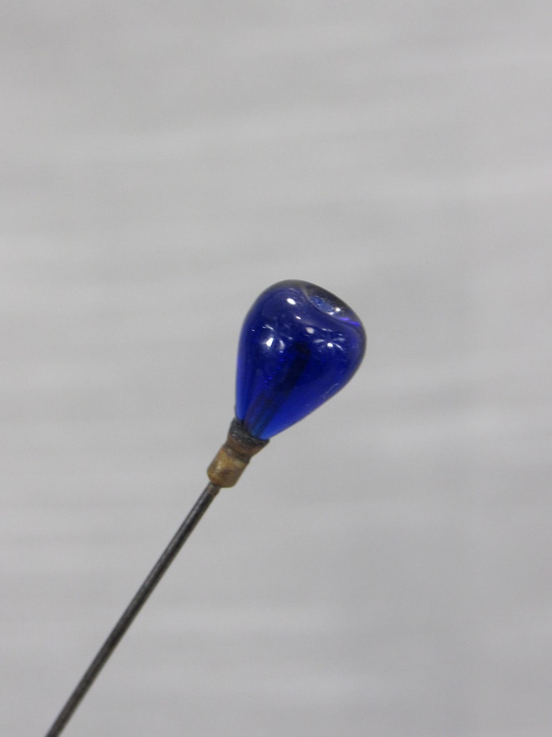 May include: A straight pin featuring a teardrop-shaped cobalt blue glass head attached to a thin metal shaft with a small brass-colored collar. The glass head has a smooth, glossy finish and is securely fastened to the pointed metal needle.
