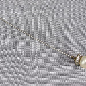 May include: A long, slender silver-toned metal lapel pin featuring a single round, off-white faux pearl at the end. The base of the pearl is accented with a small, circular metal setting embedded with tiny, clear, sparkling rhinestones.