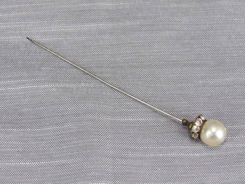 May include: A long, slender silver-toned metal lapel pin featuring a single round, off-white faux pearl at the end. The base of the pearl is accented with a small, circular metal setting embedded with tiny, clear, sparkling rhinestones.
