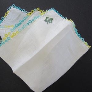 May include: White linen handkerchiefs with delicate blue and yellow lace trim. One handkerchief has a green label that reads "100% Irish Linen". The handkerchiefs are folded and displayed on a dark surface.