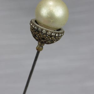 May include: A stick pin featuring a large, round, off-white pearl set within a decorative, dark metallic cup. The cup is embellished with a series of small, clear, sparkling stones arranged in a circular pattern around the base of the pearl.