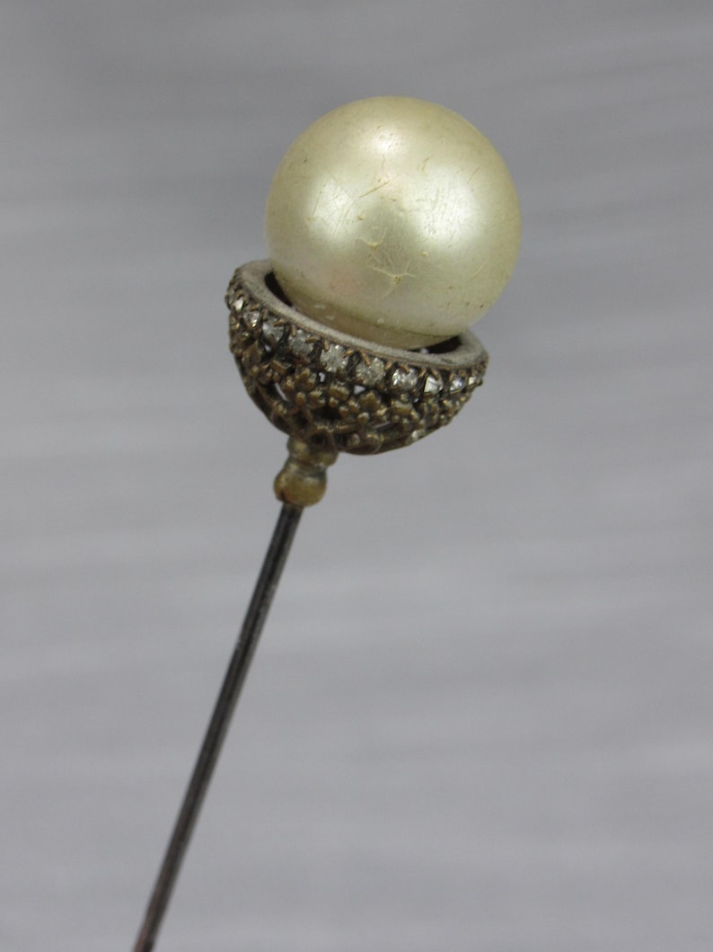 May include: A stick pin featuring a large, round, off-white pearl set within a decorative, dark metallic cup. The cup is embellished with a series of small, clear, sparkling stones arranged in a circular pattern around the base of the pearl.