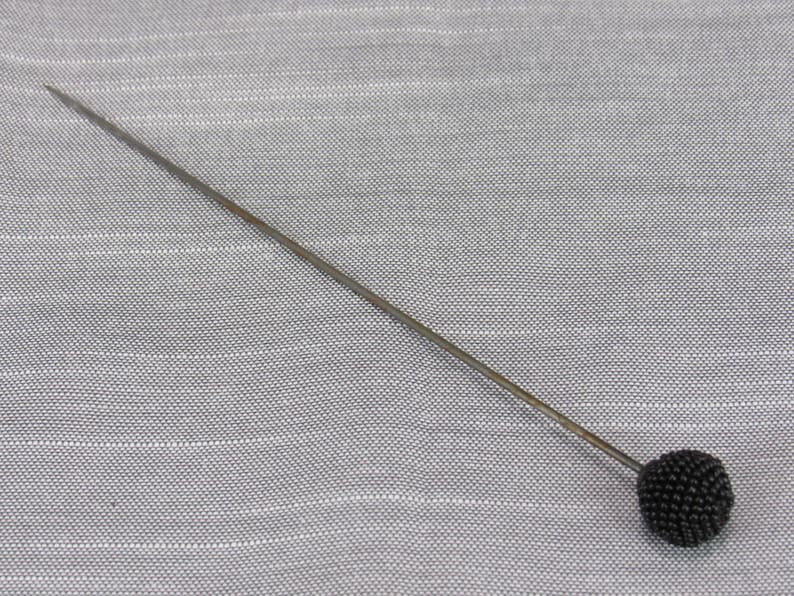 May include: A long, slender metal sewing pin featuring a round, textured black head. The pin has a sharp, pointed tip at one end and a spherical, beaded-style black head at the other, designed for securing fabric during sewing or tailoring tasks.