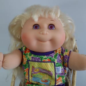 May include: A vintage Cabbage Patch Kid doll with blonde hair and purple eyes. The doll wears a colorful floral print shirt with a purple collar and sleeves. The doll has rosy cheeks and is smiling.