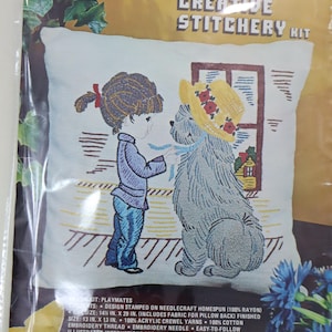 May include: Vintage "Moppets" crewel embroidery kit featuring a child and a dog wearing a hat. The kit includes fabric, yarn, needle, and instructions. Finished size is 13 in. x 13 in.