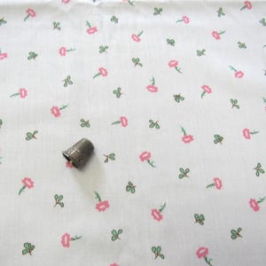 May include: A white fabric featuring a repeating pattern of small pink flowers with green leaves and stems. A metallic thimble with a textured surface is placed on the fabric to provide a sense of scale for the floral print design.