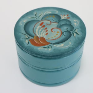 May include: A round, teal-colored lidded box featuring a hand-painted floral design on the top. The lid displays stylized petals and leaves in shades of light blue, cream, and burnt orange, set against a darker teal background.