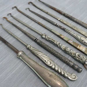 May include: A collection of six antique button hooks with metal shafts and varied decorative handles, including silver-toned metal, engraved patterns, and inlaid mother-of-pearl accents, all featuring a small curved hook at the end of each tool.