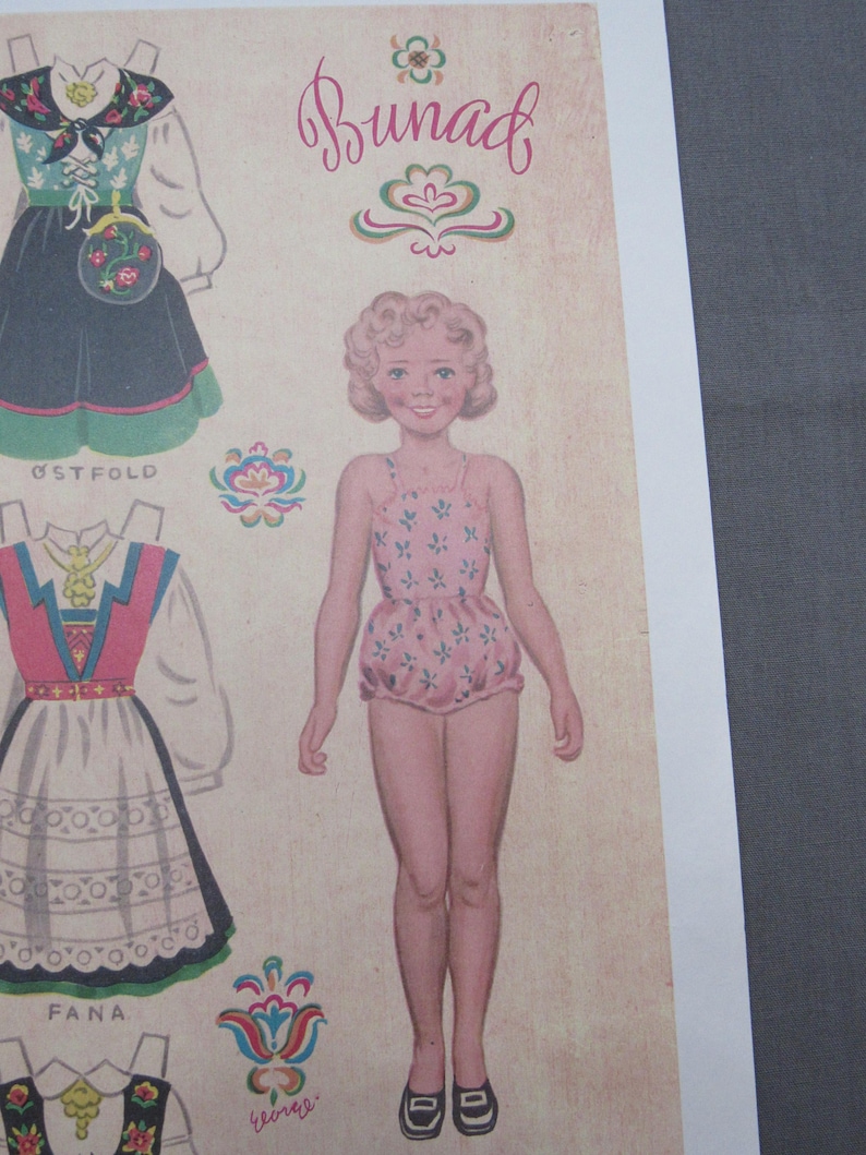Puede incluir: Hoja de mu&ntilde;eca de papel vintage con una ni&ntilde;a en un traje de ba&ntilde;o rosa floral y varios trajes folcl&oacute;ricos noruegos tradicionales. La palabra "Bunad" est&aacute; escrita en cursiva encima de la mu&ntilde;eca. Los trajes incluyen vestidos con patrones florales y geom&eacute;tricos.