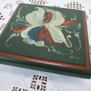 May include: A square wooden box with a dark green top and a reddish-brown frame. The top is decorated with a floral design in white, red, blue, and green. The box is resting on a white lace tablecloth. The initials "NS" and the year "1990" are visible.