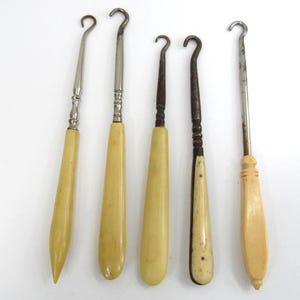May include: Five antique button hooks arranged vertically. Each tool features a curved metal hook attached to a handle. The handles vary in color and texture, ranging from smooth cream-colored material to dark, aged wood with visible grain and metal accents.