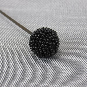 May include: A decorative hat pin featuring a spherical topper covered in small, textured black beads. The pin has a long, thin, dark metal shaft extending from the beaded sphere, which is crafted with a dense, uniform arrangement of tiny black beads.