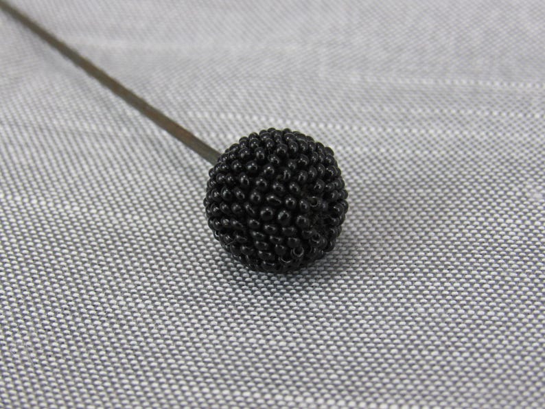 May include: A decorative hat pin featuring a spherical topper covered in small, textured black beads. The pin has a long, thin, dark metal shaft extending from the beaded sphere, which is crafted with a dense, uniform arrangement of tiny black beads.