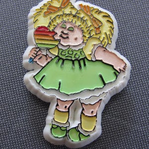 May include: A white plastic pin featuring a cartoon character with blonde hair in pigtails, wearing a light green dress and yellow shoes. The character is holding a rainbow-colored heart-shaped lollipop. The design has black outlines.