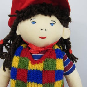 May include: A handmade doll with a red hat and brown yarn hair. The doll wears a colorful, knitted vest with red, yellow, blue, and green squares. The doll's shirt has blue and white stripes. The doll has blue eyes and a red bow.