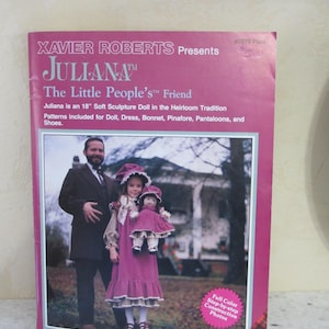 May include: A pink book cover with the title "JULIANA" and the text "The Little People's Friend." It features a photo of a man and two girls, one holding a doll. The book includes patterns for a doll, dress, bonnet, pinafore, and shoes.