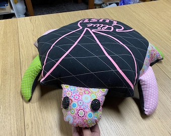 以下が含まれることがあります： ピンクと黒のキルティングの枕で、ピンクで「Cute」と書かれています。枕は亀の形をしていて、緑とピンクの生地のアクセントが付いています。