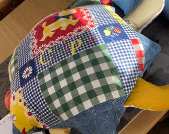 以下が含まれることがあります： 「Cuppie」と刺繍されたカラフルなパッチワークの亀の枕。デニム、格子柄、花柄など、さまざまな生地で作られています。亀は黄色、緑、赤の甲羅と黄色の腹をしています。