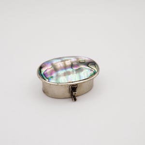 May include: An oval-shaped metal box featuring an iridescent abalone shell inlay on the lid. The box has a silver-toned finish and a small, hinged latch mechanism on the front side, allowing the lid to be opened and closed securely.