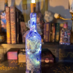 Può includere: Una bottiglia di vetro blu decorativa con un design a gufo e albero, illuminata da luci interne. La bottiglia è sormontata da un tappo di sughero marrone e una finta fiamma di candela. Circondata da libri e altri oggetti decorativi.