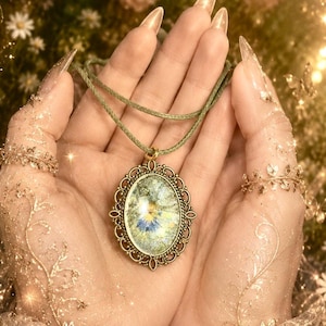 Op de afbeelding: Een ovale hangerketting met een geperste blauwe en gele bloem in een heldere hars cabochon. De hanger heeft een sierlijk, antiek ogend bronzen metalen frame met gedetailleerd filigraanwerk en hangt aan een saliegroen koord.