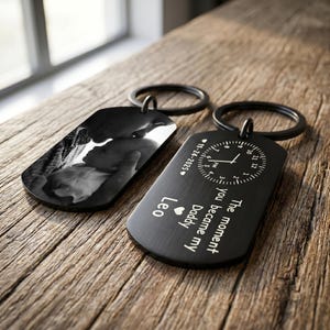 May include: Two black dog tag keychains. One features a black and white photo of a baby and a person. The other has a clock design with the text "The moment you became my Daddy Leo 11-24-2025".