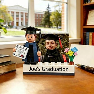 May include: A graduation-themed Lego set featuring a minifigure in a graduation cap and gown, holding a diploma. A photo of a graduate is included. The base reads "Joe's Graduation". Flowers and a pen are also visible.