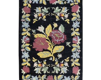 May include: A black rug with a floral pattern. The rug features a large central flower with pink and yellow petals, surrounded by smaller flowers in shades of blue, pink, and yellow. The rug has a fringed border.