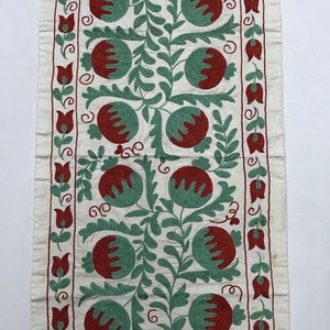 May include: A white fabric wall hanging with a repeating pattern of red and green embroidered pomegranates and leaves. The fabric has a subtle texture and the embroidery is detailed.