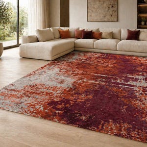 Può includere: Un grande tappeto rettangolare con un design astratto nei toni del rosso, arancione e grigio. Il tappeto è posizionato su un pavimento in legno chiaro, creando un'atmosfera calda. Il design astratto aggiunge un tocco moderno alla stanza.
