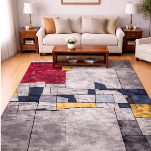 Può includere: Un tappeto moderno con un design geometrico astratto. Il tappeto presenta una base grigia con forme rettangolari rosse, blu e gialle. Il tappeto è collocato in un soggiorno con un divano, un tavolino e delle lampade.