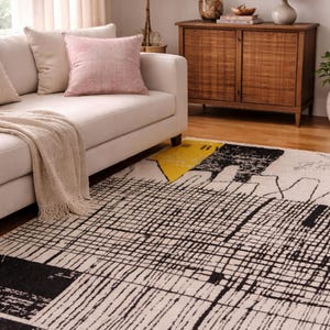 Può includere: Un tappeto moderno con un design geometrico. Il tappeto presenta uno sfondo color crema con linee e forme nere e un accento giallo. È posizionato in un soggiorno con un divano e un mobile in legno.