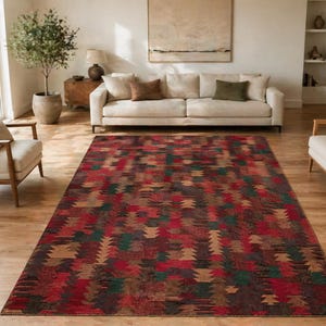 Può includere: Un tappeto rettangolare con un motivo geometrico nei toni del rosso, marrone, verde e beige. Il tappeto è posizionato su un pavimento in legno in un soggiorno, con un divano e sedie visibili sullo sfondo.