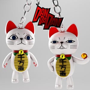 May include: Two white lucky cat keychains with red ears, blue eyes, and a gold coin-shaped pendant. One cat holds a gold ball. The keychains are attached to a silver chain with a red and black logo.