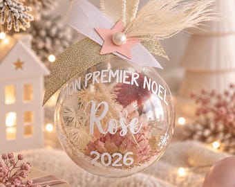Peut inclure: Une boule de Noël transparente remplie de fleurs séchées roses et blanches et de sable. L'extérieur porte l'inscription blanche MON PREMIER NOEL, Rose, et 2026. Elle est surmontée d'un ruban crème, d'un nœud doré pailleté et d'une étoile rose.