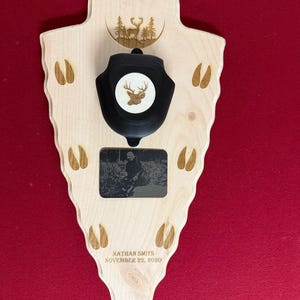 May include: A wooden arrowhead-shaped plaque with a black mounting bracket and a circular deer emblem. The plaque features a small black and white photo, deer tracks, and the engraved text "NATHAN SMITS NOVEMBER 22, 2020".